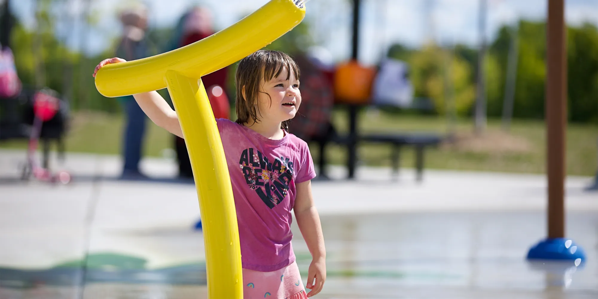 A small child stands at a colorful outdoor splash pad, holding onto a bright yellow curved water feature in the foreground. Water play structures in blue and yellow are visible around the paved area, with trees and a park setting softly blurred in the background under daylight.