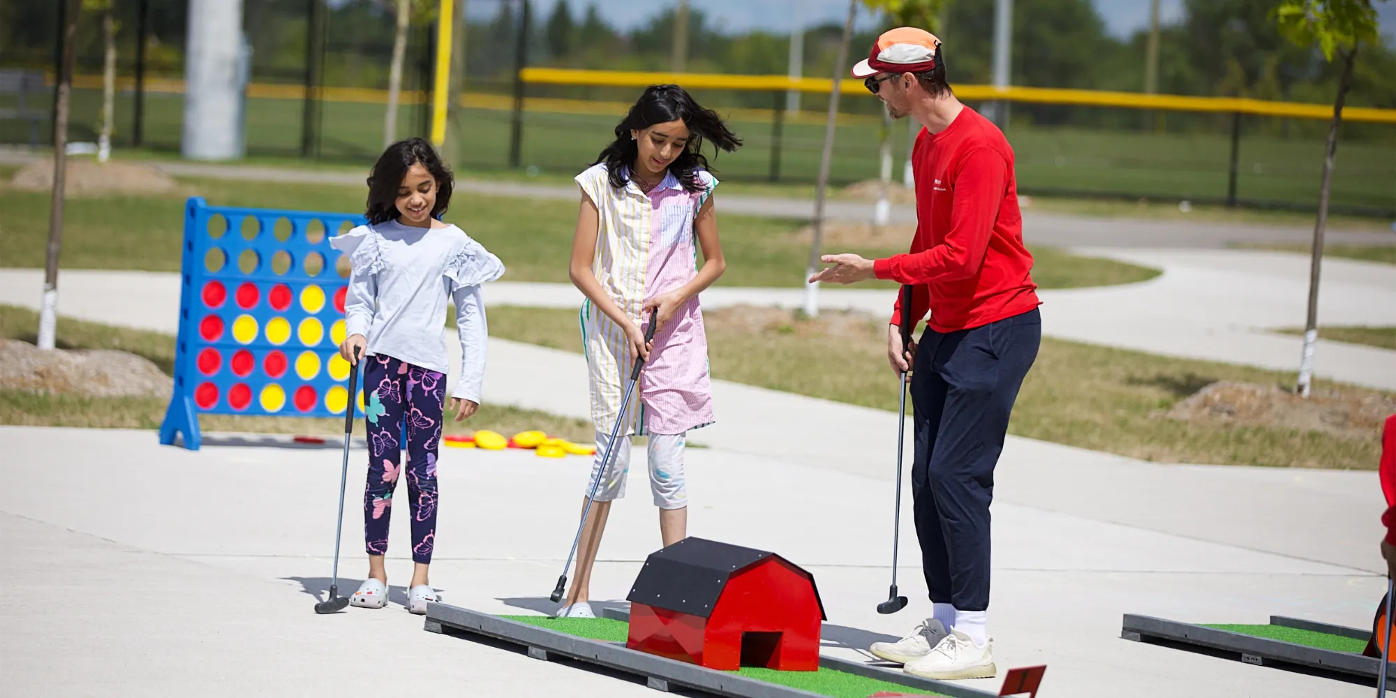 Three children and one adult stand outdoors on a paved area near a playground, ready to tee off in mini golf.