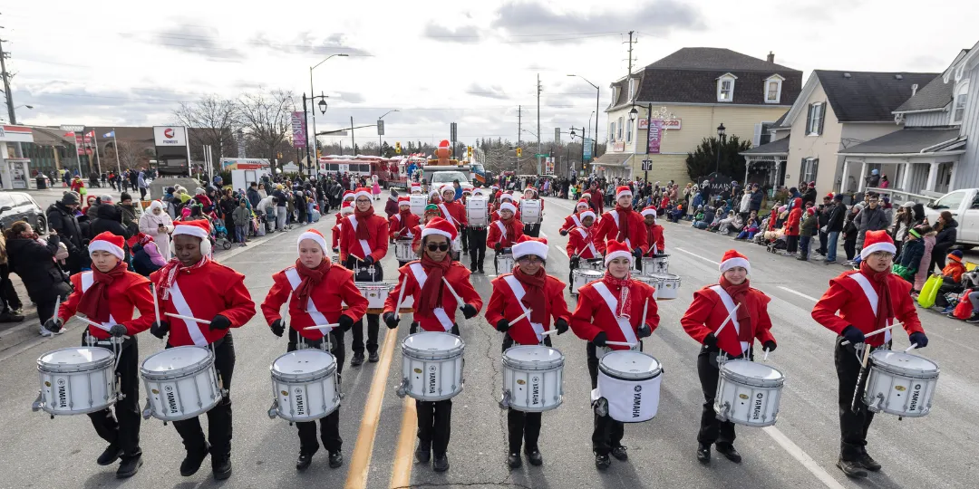 A marching band in red jackets performs outdoors on a street lined with spectators during a community parade. Residential buildings and a cloudy sky are visible in the background.